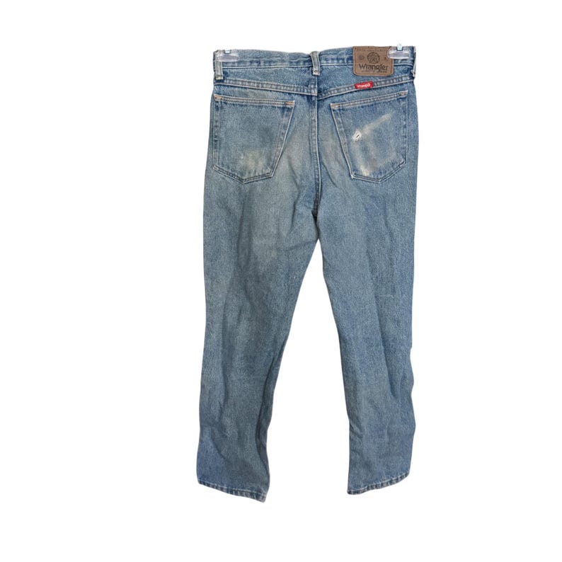 May include: Light blue denim jeans with a faded appearance. The jeans have a classic five-pocket design, a zipper fly, and a brown leather patch with the "Wrangler" logo on the back. The jeans are hanging against a white background.
