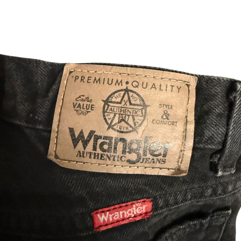 May include: Close-up of black Wrangler jeans. The jeans feature a brown leather patch with the words "Premium Quality" and "Authentic Jeans." A small red Wrangler patch is sewn on the back pocket.