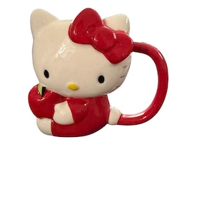 May include: A ceramic Hello Kitty mug in a seated position, with a red bow, red outfit, and white accents. The mug has a red handle and a yellow nose. The mug is a collectible item.