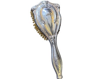 May include: An antique silver-coloured hairbrush with a decorative handle and ornate detailing. The brush head features natural bristles. The handle is engraved with floral patterns. The brush is designed with a shield-like shape.