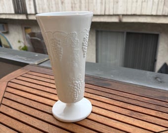 May include: A white, stemmed vase with a flared top and a round base. The vase features an embossed grape design. The vase is sitting on a wooden table.
