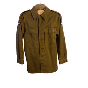Puede incluir: Chaqueta verde oliva de estilo militar con dos bolsillos en el pecho y cuello. La chaqueta tiene mangas largas y cierre de botones. Un pequeño parche con rayas rojas, blancas y azules es visible en el hombro izquierdo. La chaqueta está colgada en una percha.