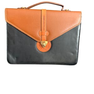 May include: A two-tone leather briefcase in black and brown. The briefcase has a flap closure with a gold-coloured clasp and a leather handle. The words "MODE ITALIANA" are visible on the strap.