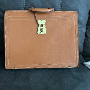 May include: A vintage brown leather briefcase with a gold-colored clasp and handle. The briefcase has white stitching around the edges and is rectangular in shape. The handle is made of brown leather.