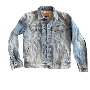 Könnte beinhalten: Hellblaue Jeansjacke mit klassischem Design. Sie hat eine Knopfleiste, zwei Brusttaschen, zwei Seitentaschen und einen braunen Lederaufnäher am Kragen. Die Jacke hat einen verblichenen, abgenutzten Look.