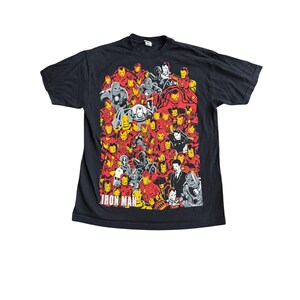 May include: A black short-sleeve t-shirt featuring a large graphic print of numerous Iron Man armored suits in red, yellow, and grey tones. The text IRON MAN is printed in white capital letters at the bottom left of the graphic design.