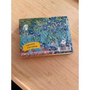 May include: A rectangular box of two decks of playing cards featuring Vincent van Gogh's Irises painting. The box displays blue irises on a green background, a yellow label with the text VINCENT VAN GOGH, and a barcode with the text 2x55 Playing Cards.
