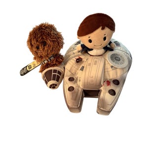 Op de afbeelding: Pluche speelgoed met personages uit Star Wars. Eén speelgoed is een bruine Chewbacca met een boogschutter, en de andere is Han Solo die op een Millennium Falcon-pluche zit. De Millennium Falcon is beige met grijze en rode details.