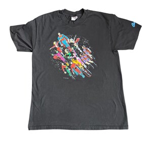 May include: A black t-shirt with a vibrant graphic of several superheroes in action. The design showcases figures in red, blue, green, and yellow, with white dots. The shirt has a crew neck and short sleeves.