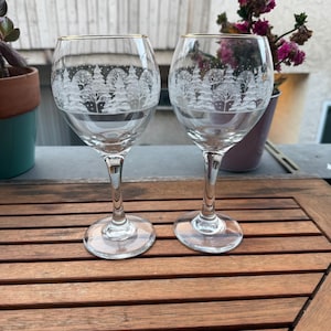 May include: Two clear wine glasses with a gold rim. Each glass has a winter forest scene etched into the glass. The glasses are on a wooden surface.