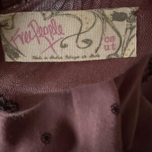 Puede incluir: Una etiqueta de ropa de la marca Free People de color morado con el texto "Free People" y "Made in India, Trimmed in Chile" y el n&uacute;mero "09 ut" en la etiqueta.