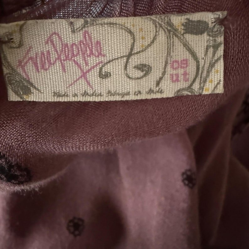Puede incluir: Una etiqueta de ropa de la marca Free People de color morado con el texto "Free People" y "Made in India, Trimmed in Chile" y el n&uacute;mero "09 ut" en la etiqueta.