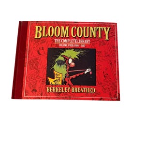 Op de afbeelding: Een rode hardcover boek met de titel "Bloom County: The Complete Library, Volume Four 1986-1987" van Berkeley Breathed. De omslag toont een cartoon illustratie van een personage met groen haar en een rode outfit. De rug is ook rood.