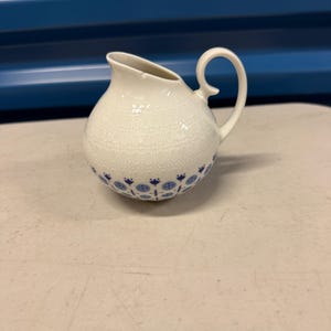 May include: A white ceramic creamer with a rounded body and a curved handle. The creamer features a delicate, textured pattern and is adorned with blue floral designs around the base. The spout is angled for easy pouring.