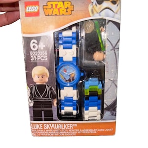 Könnte beinhalten: Eine LEGO Star Wars Luke Skywalker Armbanduhr zum Zusammenbauen mit blau-weißem Armband. Das Zifferblatt zeigt ein Star Wars-Design. Die Verpackung enthält eine Luke Skywalker Minifigur und ein grünes Lichtschwert.