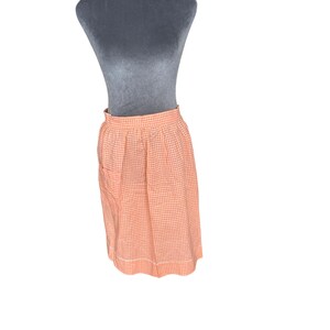 May include: A peach and white gingham skirt displayed on a mannequin. The skirt has a high waist, a pocket on the left side, and a white hemline. The fabric is a classic checkered pattern, suitable for casual wear.
