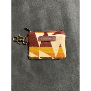 Peut inclure: Un porte-monnaie rectangulaire avec un motif géométrique dans des tons bordeaux, jaune moutarde, orange et crème. La pochette présente un écusson en cuir marron gravé du mot PENDLETON, une fermeture éclair en métal et un anneau porte-clés en laiton.