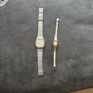 May include: Two vintage wristwatches. One is a larger, rectangular watch with a silver band and a gold-toned face. The other is a smaller, more delicate watch with a gold face and a silver band. Both watches are displayed on a dark gray surface.