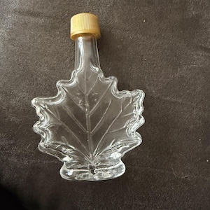 Pode incluir: Garrafa de vidro transparente em forma de folha de bordo, com uma tampa castanha clara e canelada. A garrafa, projetada para xarope ou produtos similares, tem um design detalhado de folha. Está vazia e repousa sobre um fundo escuro.