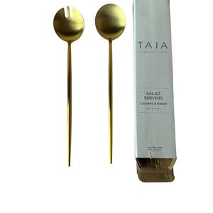 Könnte beinhalten: Zwei goldfarbene Salatbestecke mit langen, schlanken Griffen und runden Servierköpfen. Ein Besteck hat ein geschlitztes Design. Das Besteck befindet sich neben einer weißen Schachtel mit dem Text "TAJA COLLECTION" und "SALAD SERVERS".