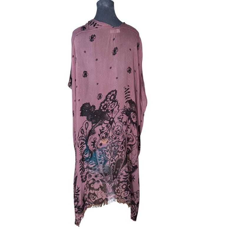 Puede incluir: Una t&uacute;nica larga, rosa y transparente, con estampado floral negro. La t&uacute;nica tiene un dobladillo con flecos y un corte holgado y fluido.
