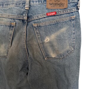 May include: Close-up of light blue denim jeans with a worn appearance. The back pockets and waistband are visible, with the "Wrangler" brand label and a red logo tag. The jeans show signs of wear and a small hole.