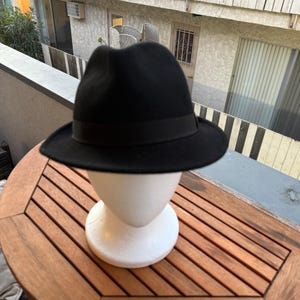 Peut inclure: Un chapeau fedora noir uni doté d'un ruban en gros-grain noir assorti autour de la calotte. Le chapeau est présenté sur une tête de mannequin blanche, mettant en valeur un design classique à calotte pincée et un bord structuré légèrement relevé.