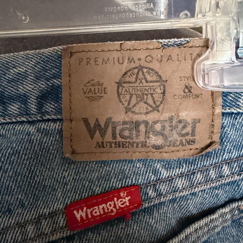 May include: Close-up of Wrangler Authentic Jeans. The jeans feature a brown leather patch with the Wrangler logo and text, including "Premium Quality" and "Authentic Jeans." A small red tag with the Wrangler logo is also visible.