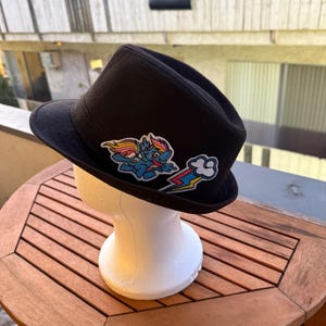 May include: A black fedora hat with two colorful embroidered patches. One patch features a blue cartoon pony with a rainbow mane and wings. The other patch shows a rainbow and lightning bolt.