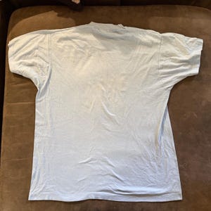 Può includere: Una t-shirt a maniche corte di colore azzurro con scollo rotondo. Il capo è steso in piano e presenta una struttura in tessuto tinta unita con cuciture visibili lungo le maniche, le spalle e l'orlo, caratterizzato da un design semplice.