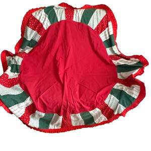 Puede incluir: Una falda redonda para árbol de Navidad con un centro rojo y un borde estampado. El borde presenta secciones alternas de lunares rojos, lunares verdes y blanco con lunares rojos. El borde está adornado con un volante rojo.