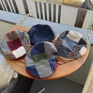 May include: Four patchwork newsboy caps in various colors and patterns are arranged on a wooden table. The caps feature a mix of fabrics in shades of blue, brown, red, and green. The caps are a classic style.