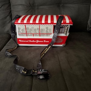 May include: Red and white striped Universal Studios Glamor Tram popcorn bucket with a black strap featuring the Universal Studios Hollywood logo.