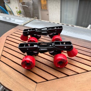 May include: Two pairs of black inline skate frames with red wheels and red toe stops are displayed on a wooden table. The wheels have a black graphic design. The frames are made of black plastic.