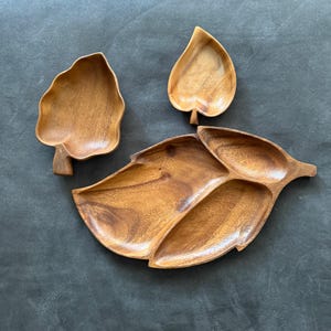 Pode incluir: Um conjunto de travessas de madeira em forma de folha. O conjunto inclui três travessas: dois pratos menores em forma de folha e uma travessa maior dividida. As travessas são feitas de madeira castanha clara com padrões de grão visíveis, perfeitas para servir petiscos.