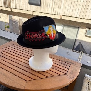 May include: Black fedora hat with a patch featuring a blonde woman and the words "THE EVIL HORDE" in red lettering. The hat is displayed on a white mannequin head.