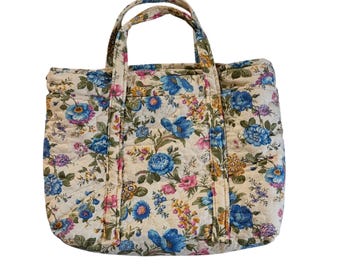 Puede incluir: Bolso tote acolchado de color crema con un estampado floral de flores azules, rosas y amarillas. El bolso tiene dos asas y forma rectangular. El diseño floral se extiende por todo el bolso.