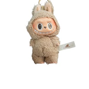 May include: A plush keychain featuring a cartoon character in a furry beige outfit with a hood and bunny ears. The character has a smiling face with large eyes and a tag that reads "THE MONSTERS".