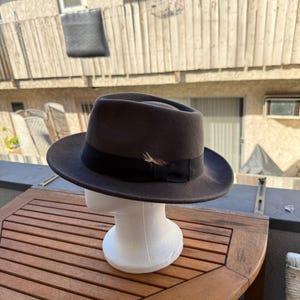 May include: A dark brown fedora hat with a black band and a small feather accent. The hat is displayed on a white mannequin head. The hat has a classic, timeless design.