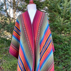 K&ouml;nnte beinhalten: Ein farbenfrohes Poncho mit V-Ausschnitt, pr&auml;sentiert auf einer Schaufensterpuppe. Das Poncho weist ein gestreiftes Muster in Rot-, Orange-, Gelb-, Blau- und Gr&uuml;nt&ouml;nen auf. Dekorative Verzierungen mit geometrischen Mustern akzentuieren die Kanten, wodurch ein lebendiges Kleidungsst&uuml;ck entsteht.
