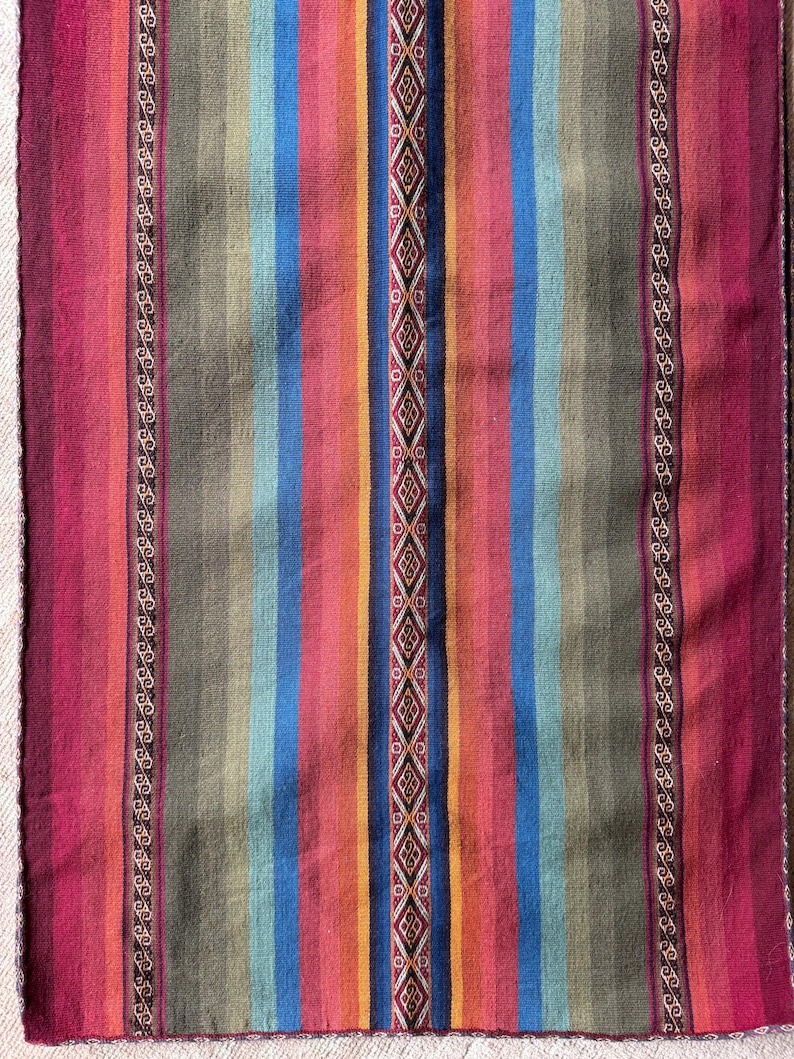 K&ouml;nnte beinhalten: Ein farbenfrohes gewebtes Textil mit vertikalen Streifen in Rot, Blau, Orange und Gr&uuml;n. Ein zentrales Rautenmuster und dekorative Bord&uuml;ren erg&auml;nzen das Design. Das Textil ist wahrscheinlich ein Teppich oder eine Decke, etwa 150 cm lang.