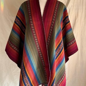 K&ouml;nnte beinhalten: Ein mehrfarbiger gestreifter Poncho auf einer Schaufensterpuppe. Der Poncho zeigt eine lebendige Mischung aus Rot, Orange, Gelb, Gr&uuml;n, Blau und Lila. Das Design beinhaltet geometrische Muster und dekorative Verzierungen.