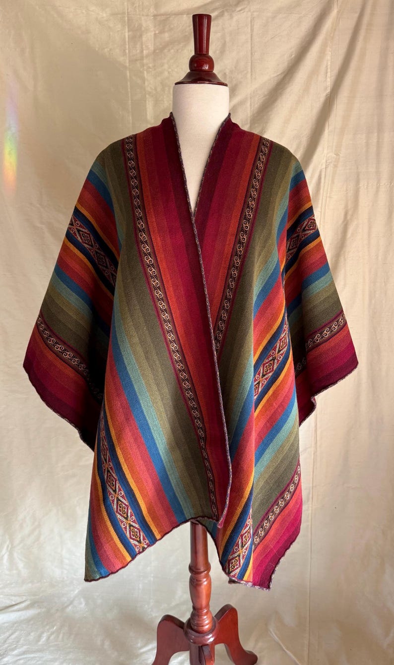 K&ouml;nnte beinhalten: Ein mehrfarbiger gestreifter Poncho auf einer Schaufensterpuppe. Der Poncho zeigt eine lebendige Mischung aus Rot, Orange, Gelb, Gr&uuml;n, Blau und Lila. Das Design beinhaltet geometrische Muster und dekorative Verzierungen.