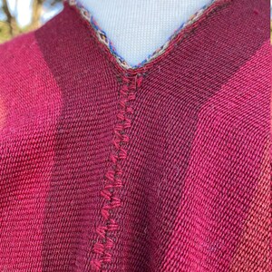 K&ouml;nnte beinhalten: Nahaufnahme eines weinroten Ponchos mit V-Ausschnitt. Der Poncho hat ein gestreiftes Muster in Rot- und Rosat&ouml;nen. Der Halsausschnitt ist mit einer bunten, gemusterten Bord&uuml;re und einer dekorativen Naht versehen.
