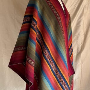 K&ouml;nnte beinhalten: Ein farbenfrohes Poncho mit einem gestreiften Muster in Rot-, Orange-, Gelb-, Gr&uuml;n- und Blaut&ouml;nen. Der Poncho wird auf einer Schaufensterpuppe pr&auml;sentiert und weist dekorative geometrische Muster an den R&auml;ndern auf. Das Kleidungsst&uuml;ck besteht aus einem weichen, gewebten Stoff.
