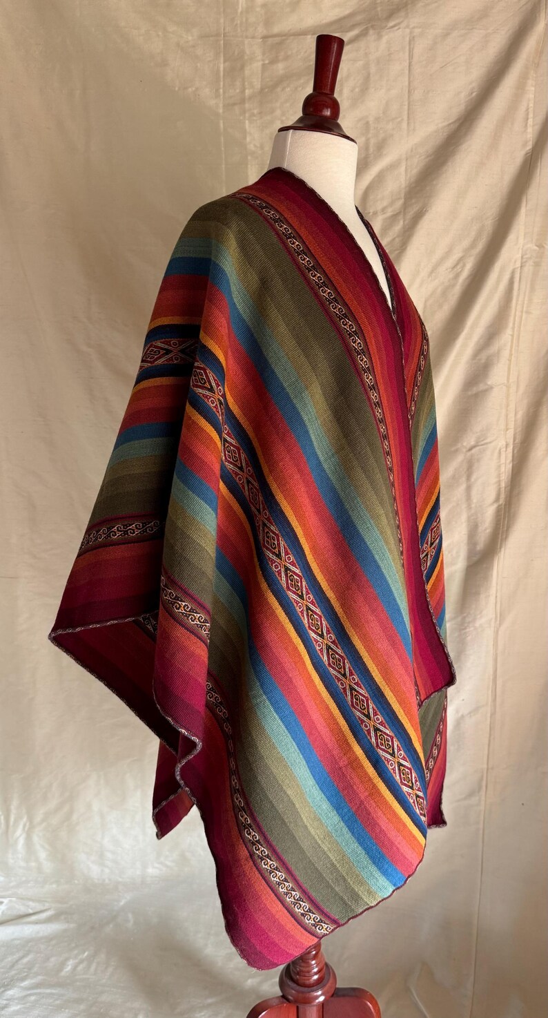 K&ouml;nnte beinhalten: Ein farbenfrohes Poncho mit einem gestreiften Muster in Rot-, Orange-, Gelb-, Gr&uuml;n- und Blaut&ouml;nen. Der Poncho wird auf einer Schaufensterpuppe pr&auml;sentiert und weist dekorative geometrische Muster an den R&auml;ndern auf. Das Kleidungsst&uuml;ck besteht aus einem weichen, gewebten Stoff.