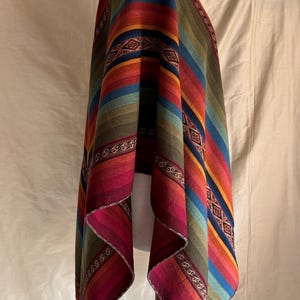 K&ouml;nnte beinhalten: Ein farbenfrohes Poncho mit horizontalen Streifen in Rot-, Orange-, Gelb-, Gr&uuml;n-, Blau- und Lilat&ouml;nen. Der Poncho wird auf einer Holzpuppe pr&auml;sentiert. Die Kanten haben dekorative Muster.