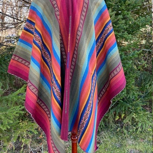 K&ouml;nnte beinhalten: Ein farbenfrohes Poncho mit Streifenmuster in Rot-, Gr&uuml;n-, Blau- und Oranget&ouml;nen. Der Poncho wird auf einem Holzst&auml;nder pr&auml;sentiert, der sein lebendiges Design und seine aufwendigen Details hervorhebt. Das Kleidungsst&uuml;ck hat einen V-Ausschnitt und dekorative Verzierungen.