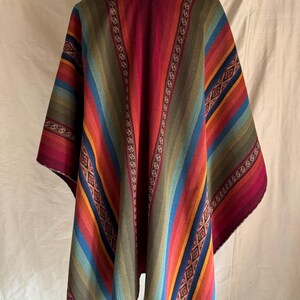 K&ouml;nnte beinhalten: Ein farbenfrohes Poncho mit Streifenmuster in Rot-, Blau-, Gr&uuml;n- und Oranget&ouml;nen. Der Poncho wird auf einer Holzpuppe pr&auml;sentiert. Das Design umfasst geometrische Muster und dekorative Bord&uuml;ren.