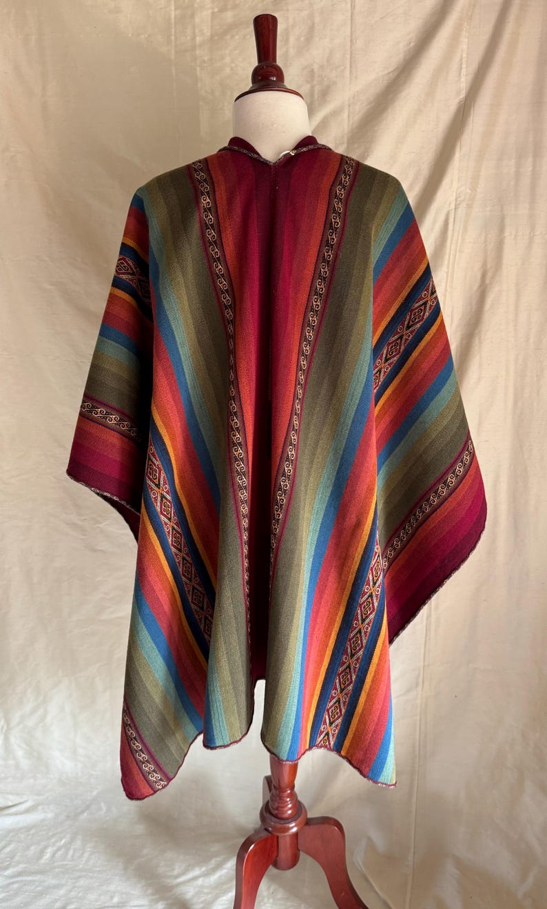 K&ouml;nnte beinhalten: Ein farbenfrohes Poncho mit Streifenmuster in Rot-, Blau-, Gr&uuml;n- und Oranget&ouml;nen. Der Poncho wird auf einer Holzpuppe pr&auml;sentiert. Das Design umfasst geometrische Muster und dekorative Bord&uuml;ren.
